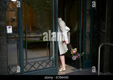 Donna con un cappotto grigio che entra in un edificio che tiene fiori, vista da dietro. Bruxelles, Belgio Foto Stock