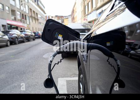 Auto elettrica collegata a una stazione di ricarica su una strada della città, circondata da edifici e auto parcheggiate. Germania Foto Stock