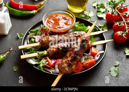 Kebab lula preparato su spiedini servito con verdure fresche e salsa di pomodoro su piatto nero su sfondo scuro. Vista ravvicinata Foto Stock