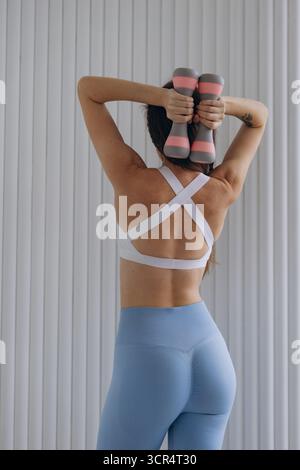 Vista posteriore corpo atletico giovane donna con leggings blu reggiseno sportivo bianco che si allenano manubri in interni. Corpo tonificato, fitness e stile di vita sano Foto Stock