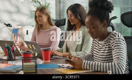 Tre donne d'affari informali multirazziali che lavorano in ufficio donne d'affari informali che lavorano con computer portatili scrivono appunti sorridenti guardando Foto Stock