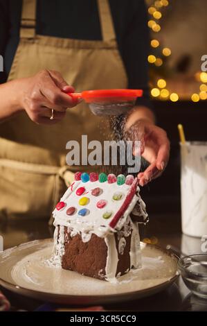 Una persona all'interno di un grembiule spolvera di zucchero in polvere su una casa festosa di pan di zenzero in una cucina calda e illuminata. Foto Stock