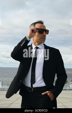 Uomo di mezza età caucasico che indossa tuta e occhiali da sole all'aperto vicino al lungomare che regola gli occhiali da sole con un'espressione sicura che guarda lontano dalla fotocamera Foto Stock