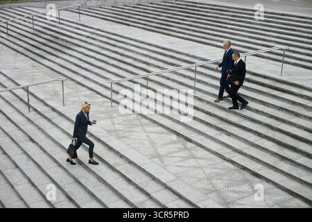 Una giovane donna d'affari caucasica che salta i gradini all'aperto tenendo in mano caffè e borsa, mentre due uomini in tute formali scendono in direzione opposta, lasciando dopo le trattative Foto Stock