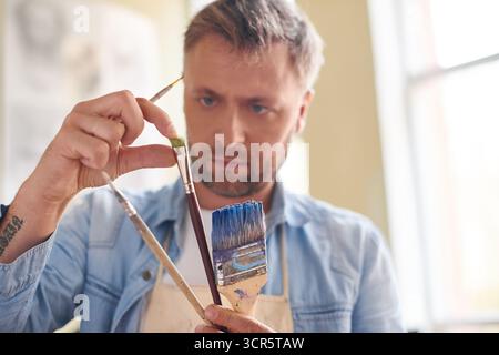 Preparatevi per la pittura. Artista concentrato con la barba che tiene in mano pennelli di diverse dimensioni e ne controlla la qualità Foto Stock
