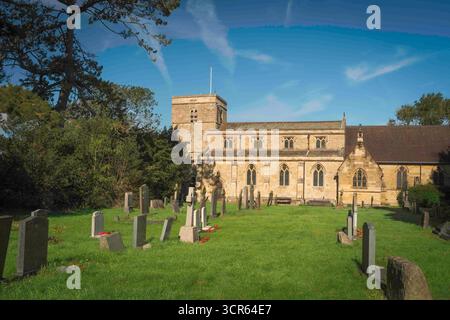 Chiesa di tutti i Santi nella splendida parrocchia locale sotto un cielo luminoso con razze di nuvole e fiancheggiata da alberi e prati consolidati a Bishop Burton, Regno Unito. Foto Stock