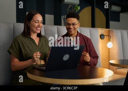 Giovani uomini d'affari allegri in smart casuals che celebrano il successo mentre utilizzano il laptop nella sala dell'ufficio Foto Stock