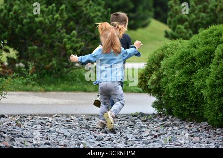 Bambina dai capelli rossi con trecce che inseguono ragazzo con una camicia nera sul sentiero di Gravel Park. I bambini che corrono, ridono e si divertono all'aria aperta senza pensieri Foto Stock