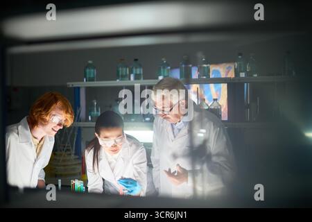 Scienziati in occhiali protettivi che discutono i risultati della ricerca in laboratorio. Ricercatrice asiatica donna che tiene provetta con liquido rosso in aveva mentre i suoi colleghi maschi che lo guardano con fiducia Foto Stock