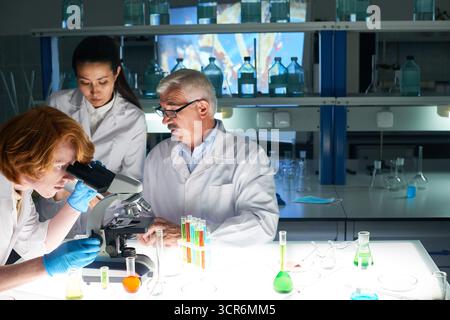 Biochimici che effettuano esperimenti e annotano i risultati negli appunti. Giovane assistente con i capelli di zenzero che guarda attraverso il microscopio e il suo collega anziano seduto al tavolo e che si occupa della gestione Foto Stock