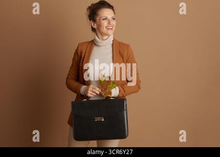 Una donna d'affari sorridente con dolcevita beige e blazer marrone guarda lateralmente, tenendo in mano una valigetta in pelle nera e un gruppo di foglie autunnali Foto Stock