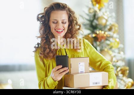 Una donna con lunghi capelli ricci ed espressione allegra può contenere smartphone e piccole scatole di cartone. Con una camicetta gialla brillante, si erge davanti a una luce soffusa Foto Stock
