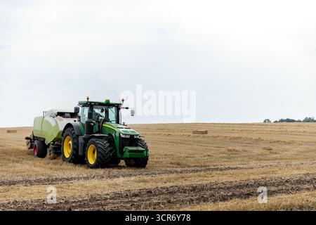 Un potente trattore verde gestisce i raccolti sul campo, raccogliendo fieno durante l'attività agricola, circondato da ampi campi sotto un cielo luminoso con soffici nuvole Foto Stock