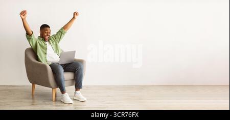 Joyful African Male Using laptop shaking Fists over Grey Wall Foto Stock