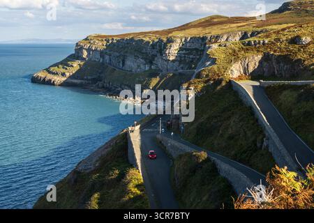 Questa suggestiva fotografia costiera mostra una strada tortuosa scavata in scogliere spettacolari, con un'auto rossa che guida lungo il percorso che si affaccia sul profondo blu Foto Stock