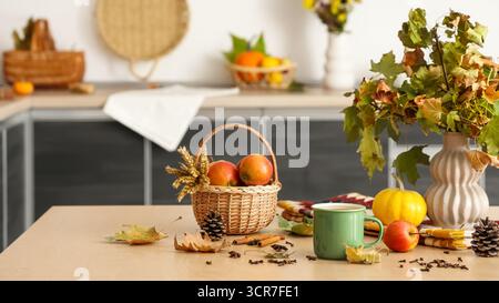 Vaso con foglie autunnali, tazza di tè caldo, cestino con mele e spezie sul tavolo in cucina Foto Stock