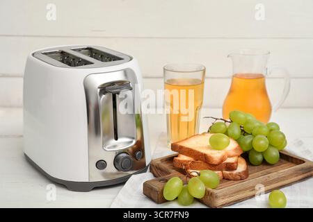 Moderno tostapane con fette di pane e uva su sfondo di legno bianco Foto Stock