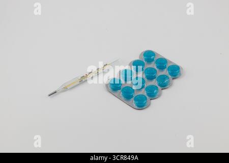 Le capsule blu brillante per medicinali sono disposte in un vassoio accanto a un termometro in vetro su una superficie bianca semplice. Questa configurazione suggerisce salute e benessere. Foto Stock