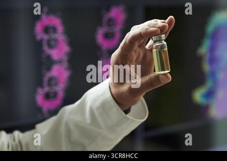 Mano di un medico di sesso maschile in camice bianco da laboratorio contenente fiala trasparente con liquido medico giallo in ambiente conferenza, dati di ricerca scientifica e display digitali colorati sullo sfondo Foto Stock
