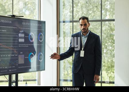 Giovane ricercatore maschio che presenta statistiche mediche su grande schermo, in piedi in ambiente ufficio moderno, tenendo puntatore, indossando abito da lavoro, guardando il pubblico Foto Stock
