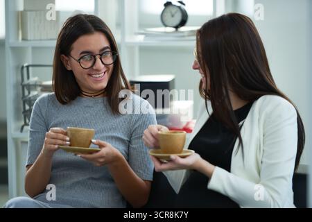 Giovane donna elegante con taglio a Bob che indossa un abito grigio e occhiali rotondi seduti sul divano con la sua collega. Parlano e sorridono allegramente mentre bevono caffè da piccole tazze beige Foto Stock