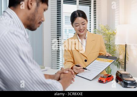 Una negoziazione per la vendita di auto si svolge in un ufficio in cui una donna venditrice con un blazer giallo spiega i dettagli di un contratto a un cliente maschio, showin Foto Stock