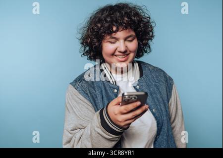 Giovane con capelli ricci che coinvolge lo smartphone su uno sfondo blu in un ambiente informale Foto Stock