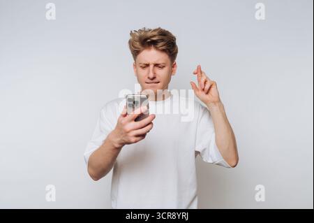 Il giovane controlla il telefono con un'espressione di speranza mentre incrocia le dita per avere fortuna in un ambiente luminoso e minimalista Foto Stock