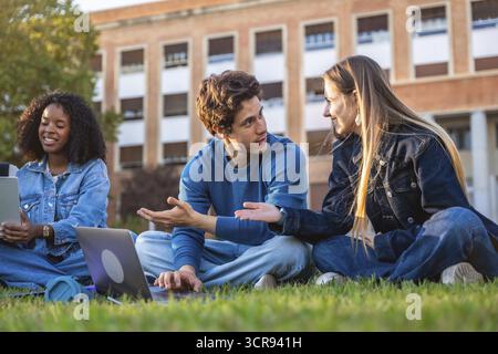 Gruppo di giovani studenti universitari seduti sull'erba verde che parlano e studiano casualmente con i computer portatili, trascorrendo un caldo pomeriggio al campus Foto Stock