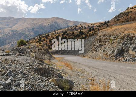 Veduta di una strada tortuosa che attraversa il paesaggio arido e roccioso, salendo in lontane montagne sotto un cielo di nuvole sparse, passo dei Vardenyats, Armenia. Foto Stock