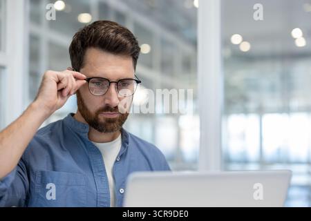 Uomo d'affari concentrato che regola gli occhiali mentre lavora intensamente su un notebook in un ufficio moderno, creando un senso di concentrazione, professionalità e dedizione alle sue attività Foto Stock
