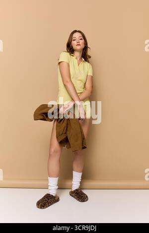 Una giovane donna indossa con sicurezza un elegante abito giallo e una giacca marrone sullo sfondo beige. Foto Stock
