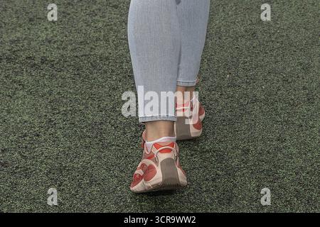 Primo piano di gambe femminili in sneakers rosse e jeans blu che camminano su un terreno sportivo verde. Concetto di stile di vita attivo, fitness, moda informale e curativo Foto Stock