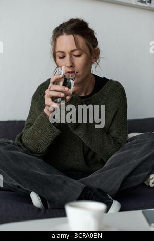 La donna si siede a gambe incrociate sul divano, perduta nel pensiero mentre beveva acqua in silenzio. Foto Stock