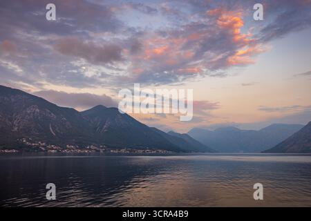 Tramonta la luna nella calma pre-alba sopra la baia di Cattaro, in Montenegro, con montagne nebbiose, nuvole morbide e cielo blu-rosa che si riflette nelle acque tranquille prima dell'alba Foto Stock