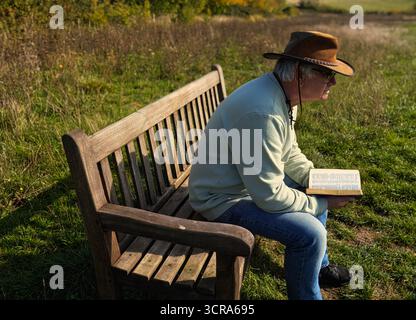 Uomo anziano che indossa un cappello di cuoio che legge un libro sulla panchina di un parco al caldo sole della sera contemplando la vita Foto Stock