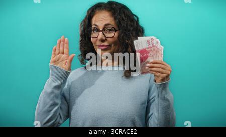 Donna di mezza età che tiene in mano banconote della corona islandese mentre alza la mano con un gesto di giuramento su uno sfondo blu. Foto Stock