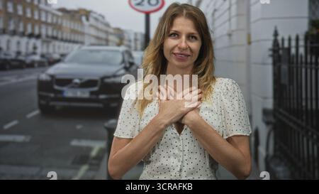 Una donna con un abito bianco a pois mette le mani sul petto in un gesto grato su una strada della città con auto parcheggiata e un cartello visibile a 20 mph; gratitudine heali Foto Stock