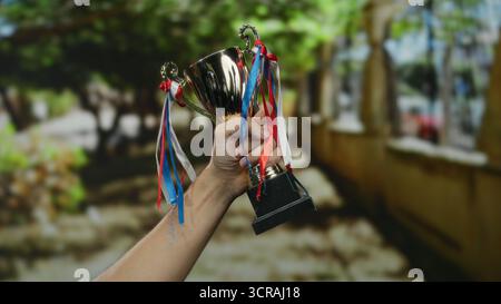 Mano di un uomo caucasico che tiene il trofeo in città all'aperto celebrando la vittoria con nastri blu rossi bianchi. Foto Stock