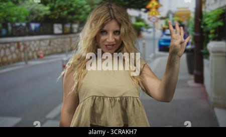 Donna che saluta la mano per strada con capelli biondi e top beige senza maniche, sorride e prepara un saluto a palma aperta alla fotocamera; un saluto amichevole. Foto Stock