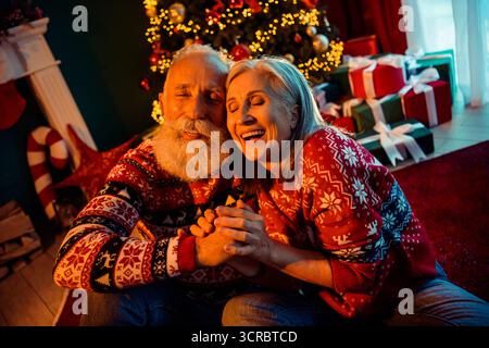 Una coppia in pensione gioiosa condivide un caldo momento natalizio a casa con un albero decorato regali maglioni accoglienti e luci festive che creano una vacanza amorevole Foto Stock