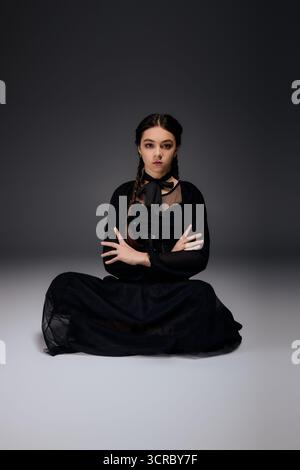 Una ragazza con trecce sede a gambe incrociate, vestita con un abito gotico Black Wednesday per celebrare Halloween. Foto Stock