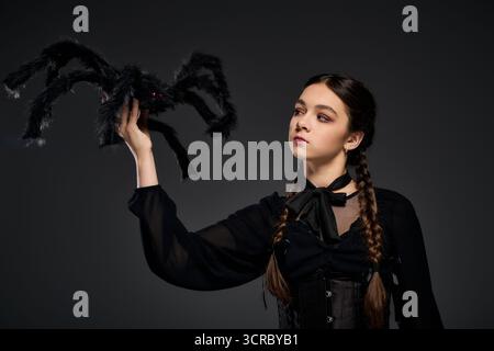 Una ragazza con trecce indossa un costume gotico del mercoledì, tenendo in mano un ragno spaventoso in un ambiente buio. Foto Stock