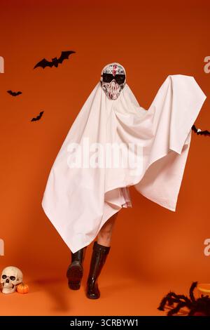 Un bambino in costume da fantasma celebra con gioia Halloween su uno sfondo arancione. Foto Stock