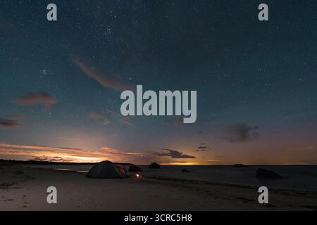 Una tenda luminosa su una tranquilla spiaggia sabbiosa sotto uno spettacolare cielo stellato, un'idilliaca scena di campeggio sul mare. Foto Stock