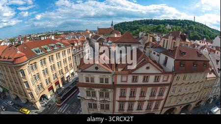 La lente fisheye cattura il fascino della città vecchia di Praga, mostrando i suoi tradizionali tetti rossi, una strada trafficata con un tram giallo e lussureggianti colline verdi Foto Stock