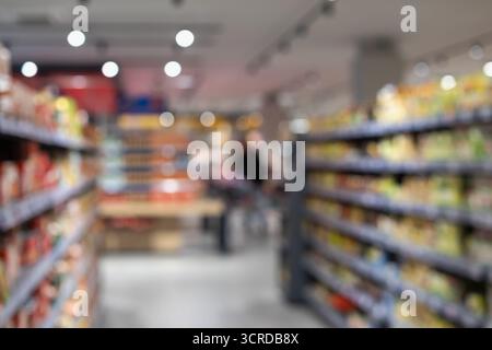 foto sfocata di un corridoio luminoso del supermercato con scaffali pieni di prodotti e un acquirente lontano con un carrello. ideale per argomenti come la comunicazione Foto Stock