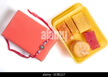Una vivace mostra che presenta una varietà di dolci indiani in una scatola gialla, accompagnata da una scatola regalo rossa decorata con un rakhi. Foto Stock