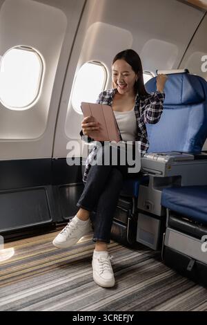 Giovane donna asiatica seduta su un sedile in aereo, sorride e punta su un tablet digitale. Sembra che stia godendo di intrattenimento o video a bordo c Foto Stock