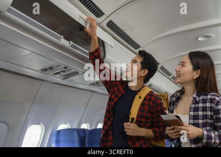 Coppia asiatica che si prepara a viaggiare su un aereo. l'uomo sta mettendo il suo oggetto nel vano superiore, mentre la donna sorride e tiene in mano passaporti e boa Foto Stock